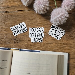 Könnte beinhalten: Drei weiße Aufkleber mit schwarzer Beschriftung auf einer Holzoberfläche. Die Aufkleber sagen "You are enough", "You can do hard things", und "See the good".