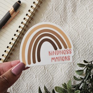 Könnte beinhalten: Ein Aufkleber mit einem Regenbogen-Design in Brauntönen und dem Text "Kindness Matters" in Rosa.