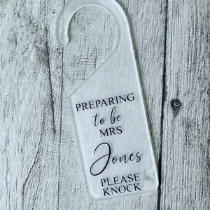 May include: A clear, frosted door hanger with a curved top. The text "PREPARING to be MRS Jones PLEASE KNOCK" is printed in black script. The background is a light grey wood grain.