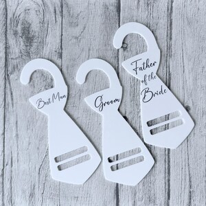 May include: Three white tie-shaped wedding hangers. Each has a curved top and two horizontal slots. The hangers are inscribed with the phrases "Best Man", "Groom", and "Father of the Bride" in black script.