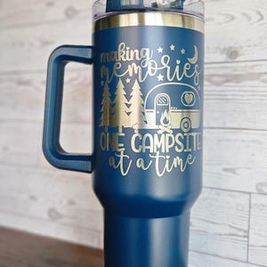 May include: A blue stainless steel travel mug with a handle and a lid. The mug has a gold inscription that reads "Making memories one campsite at a time".