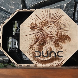Puede incluir: Caja de madera con una tapa que presenta un gráfico marrón y blanco de un gusano de arena y la palabra "DUNE". La caja contiene tres vasos de chupito transparentes con diseños grabados.