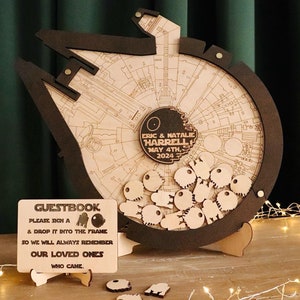 Può includere: Un libro degli ospiti in legno a forma di Millennium Falcon di Star Wars con un cartello che recita "GUESTBOOK PLEASE SIGN A & DROP IT INTO THE FRAME SO WE WILL ALWAYS REMEMBER OUR LOVED ONES WHO CAME". Il libro degli ospiti ha una cornice nera e un cerchio di legno con un disegno dell'interno del Millennium Falcon. Il cerchio ha un piccolo cerchio nero con il testo "ERIC & NATALIE HARRELL MAY 4TH, 2024" su di esso. Ci sono piccoli pezzi di legno a forma di Morte Nera nel cerchio.