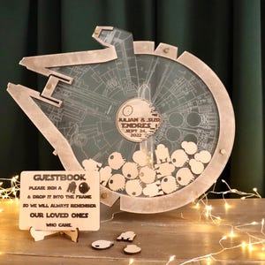 May include: A wooden Star Wars X-Wing spaceship themed wedding guest book with a clear acrylic frame. The frame is filled with small wooden cutouts of the Millennium Falcon. The sign reads "GUESTBOOK PLEASE SIGN A & DROP IT INTO THE FRAME SO WE WILL ALWAYS REMEMBER OUR LOVED ONES WHO CAME".