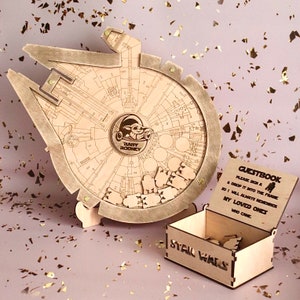 May include: A wooden Star Wars themed guest book with a Millennium Falcon design. The guest book is made of wood and has a small box with a sign that says "Stall Walls". The box is open and has small wooden pieces inside. The sign on the box says "Guestbook, Please sign a & drop it into the frame so I will always remember my loved ones who came."