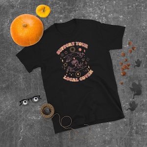 Puede incluir: Camiseta negra con un diseño gráfico de una mano con un ojo en la palma, rodeada de estrellas, lunas y el texto "Support Your Local Coven".