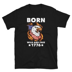 Op de afbeelding: Zwart t-shirt met een patriottisch ontwerp. De afbeelding toont een Amerikaanse zeearend met de Amerikaanse vlag, vlammen en de tekst "BORN WILD AND FREE 1776".