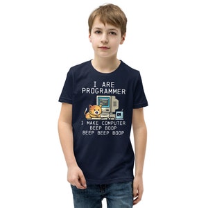 Op de afbeelding: Een donkerblauw t-shirt voor kinderen met een cartoonkat die aan een computer zit. De tekst op het shirt luidt "I are programmer I make computer beep boop beep beep boop".