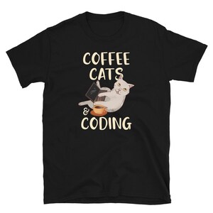 May include: Black t-shirt featuring a graphic of a white cat using a laptop with a coffee cup beside it. The text "COFFEE CATS & CODING" is printed above and below the cat illustration in a whimsical font.