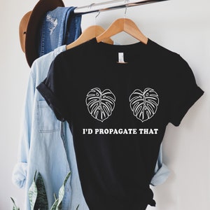 Pode incluir: T-shirt preto com gr&aacute;fico branco de duas folhas de monstera e o texto "I'D PROPAGATE THAT". A camisa est&aacute; pendurada em um cabide de madeira, com outras roupas ao fundo.
