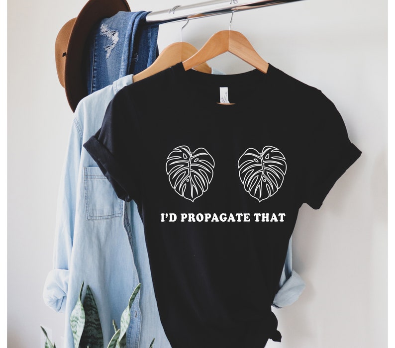 Pode incluir: T-shirt preto com gr&aacute;fico branco de duas folhas de monstera e o texto "I'D PROPAGATE THAT". A camisa est&aacute; pendurada em um cabide de madeira, com outras roupas ao fundo.