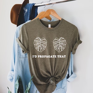 Pode incluir: Camiseta verde-oliva com desenhos de folhas de monstera brancas e o texto "I'd Propagate That". A camiseta est&aacute; pendurada em um cabide de madeira com outras roupas ao fundo.