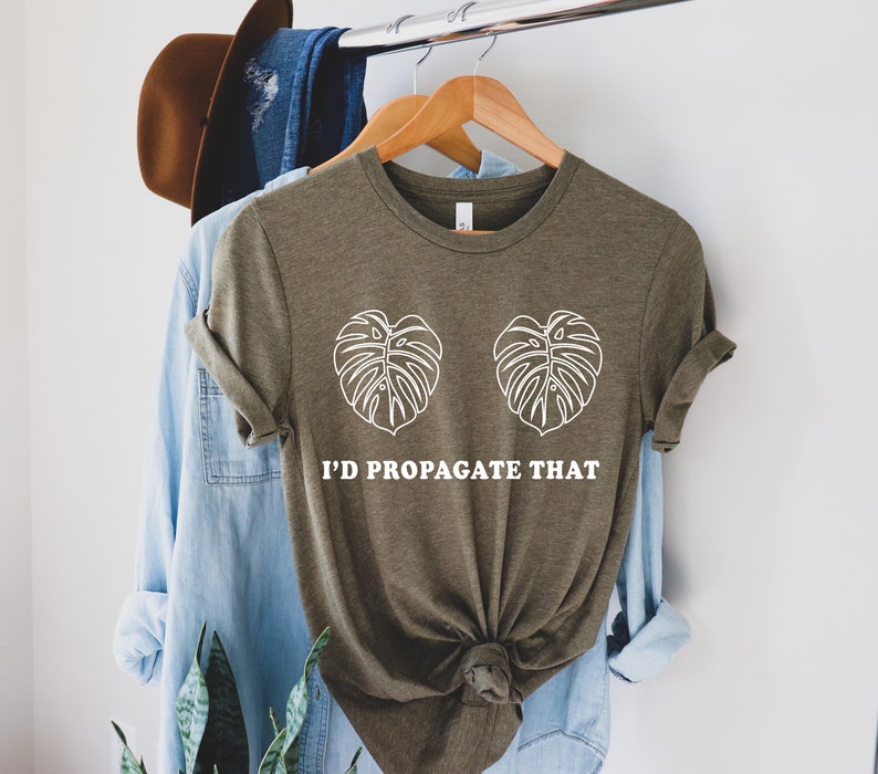 Pode incluir: Camiseta verde-oliva com desenhos de folhas de monstera brancas e o texto "I'd Propagate That". A camiseta est&aacute; pendurada em um cabide de madeira com outras roupas ao fundo.