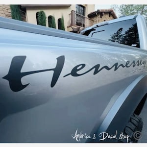 May include: A silver pickup truck with a black "Hennessey" decal on the side. The decal is in a cursive font.
