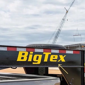 以下が含まれることがあります： 黄色い「BigTex」の文字と赤と白の反射ストライプが付いた黒いトレーラー。背景にはクレーンとスペアタイヤが見えます。トレーラーはトラックに接続されています。