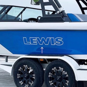 Könnte beinhalten: Ein weißes und blaues Boot mit dem Namen "LEWIS" in weißen Buchstaben auf einer blauen Tafel. Das Boot hat schwarze Reifen und Felgen. Ein Pfeil zeigt nach oben zum Namen. Das Boot steht auf einem Anhänger.