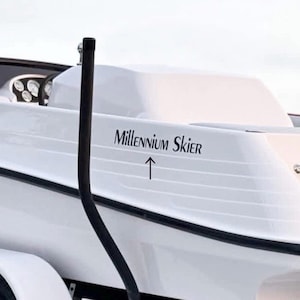 Op de afbeelding: Een witte boot met de tekst "Millennium Skier" in zwarte letters. De boot heeft een zwarte rand en een zwarte paal. De boot staat op een aanhanger.