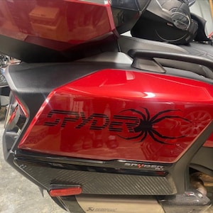 May include: A red Spyder motorcycle with a black spider graphic on the side. The motorcycle has a black seat and a black exhaust pipe.