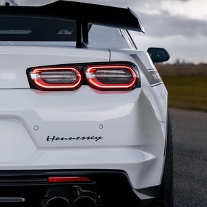 May include: Rear view of a white Hennessey-modified Chevrolet Camaro with a black rear spoiler, black racing stripes, and dual exhaust pipes.