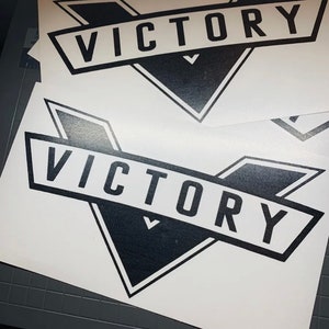 Może przedstawiać: Kilka białych naklejek winylowych z czarno-białym wzorem. Każda naklejka zawiera słowo "VICTORY" pogrubioną, bezszeryfową czcionką w czarnym geometrycznym kształcie. Naklejki znajdują się na szarej powierzchni.