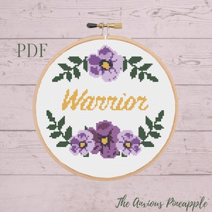 Peut inclure: Un modèle de point de croix avec une couronne de fleurs comprenant des fleurs violettes et des feuilles vertes entourant le mot "Warrior" en texte jaune. Le motif est présenté dans un cercle de broderie en bois.