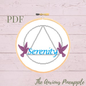 May include: A cross-stitch pattern featuring a circle with a triangle inside, two purple birds on either side of the word "Serenity" in blue text. The pattern is in a wooden embroidery hoop.