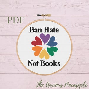 Puede incluir: Un patrón de punto de cruz con un diseño de corazón arcoíris con el texto "Ban Hate Not Books". El patrón está en un aro de bordado de madera.