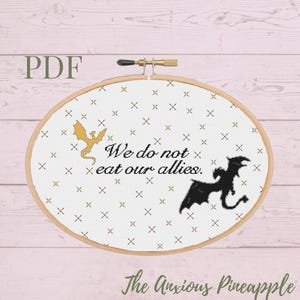 May include: A white embroidery hoop with a gold and black dragon design. The text "We do not eat our allies." is printed in black on the white fabric. The hoop is on a pink wooden background.