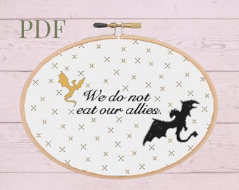 May include: A white embroidery hoop with a gold and black dragon design. The text "We do not eat our allies." is printed in black on the white fabric. The hoop is on a pink wooden background.