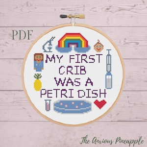 Puede incluir: Un aro de bordado blanco con un diseño de punto de cruz que dice "Mi primera cuna fue una placa de Petri". El diseño incluye un arcoíris, un bebé, una piña, un tubo de ensayo, una placa de Petri, una jeringa y un corazón.