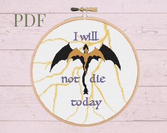 May include: A white embroidery hoop with a cross-stitch design featuring a dragon with the text "I will not die today". The hoop is on a pink wooden background.