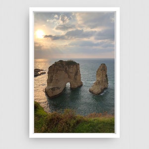 May include: A scenic photograph of two large rock formations with a natural archway in the ocean. The rocks are a light brown color and are surrounded by blue water. The sky is a light blue with white clouds and the sun is shining brightly in the distance.