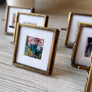 May include: Several small, square brass picture frames with miniature art prints. Each frame has a white mat and a small stand on the back. The artwork features colorful floral paintings.
