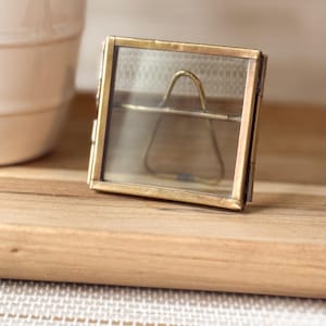 May include: A small, rectangular brass picture frame with clear glass panels. The frame has a hinged design and a small, gold-colored handle inside. The frame is displayed on a wooden surface, with a ceramic mug in the background.