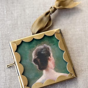 May include: A square brass frame with a scalloped edge containing a portrait of a person seen from behind. The frame features a hinged side closure and is attached to a dark olive green ribbon for hanging. The portrait shows a person with dark hair.