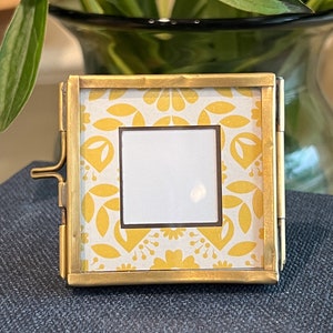 May include: A small, square, hinged picture frame with a gold-colored metal frame. The interior features a white square with a yellow and white floral pattern. The frame is open, revealing the interior design.