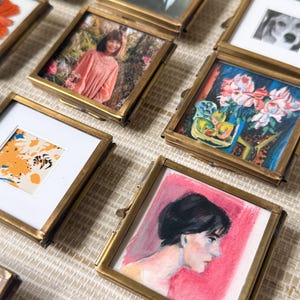 May include: A collection of small, square brass picture frames, each containing a different image. The images include a portrait, a dog, a floral still life, and abstract art. The frames are arranged on a textured surface.