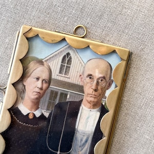 May include: A rectangular brass picture frame with a scalloped edge and a top hanging loop. The frame contains a print of the American Gothic painting, featuring a person with hair pulled back and a person with glasses holding a pitchfork.