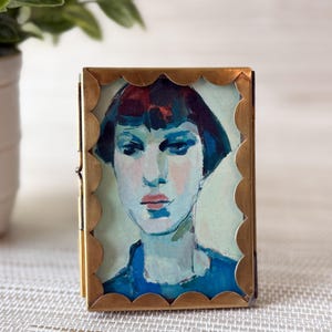 May include: A small, rectangular portrait in a gold scalloped frame. The artwork depicts a woman with blue and red hair, wearing a blue top. The painting has a soft, impressionistic style, with a focus on the subject's face.