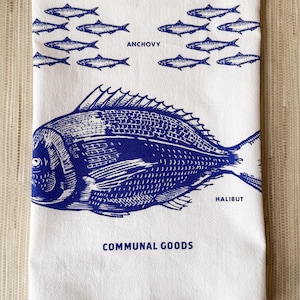 May include: White kitchen towel with blue illustrations of fish, including trout, anchovy, and halibut. The towel has the words "Communal Goods" printed at the bottom.