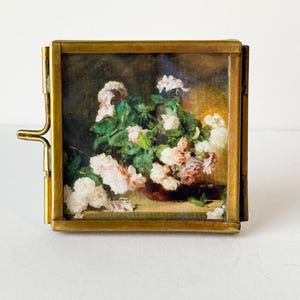 May include: A small, hinged brass frame holds a floral painting. The painting features a bouquet of pink and white roses with green foliage against a dark background. The frame has a clasp on the side.