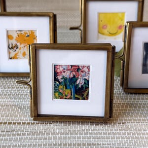 May include: A collection of small, square brass picture frames with various images. One frame displays a colorful floral print, while others showcase a smiling face, a dog, and abstract art. The frames are arranged on a textured surface.