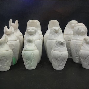 Op de afbeelding: Een set van tien witte keramische canopische potten met verschillende Egyptische goden- en dierenfiguren. De potten zijn ingewikkeld gegraveerd met hiërogliefen en hebben deksels.
