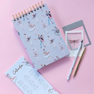 Puede incluir: Un cuaderno gris de espiral con un calendario en la portada. El cuaderno está decorado con un patrón de bailarinas en varias poses. El cuaderno está sobre una superficie rosa con un marco de fotos plateado y una libélula dentro.