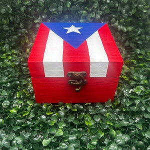 May include: A red wooden box with a hinged lid, painted with the Puerto Rican flag design. The top features a blue triangle with a white star, and alternating white and red stripes. The box has a bronze-colored clasp.