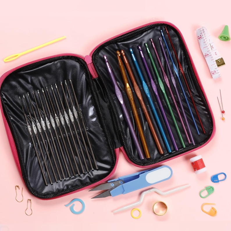 May include: A pink zippered crochet hook case open to reveal a variety of crochet hooks in different colors and sizes. Additional tools include scissors, stitch markers, a measuring tape, and yarn needles. The tools are arranged on a pink surface.