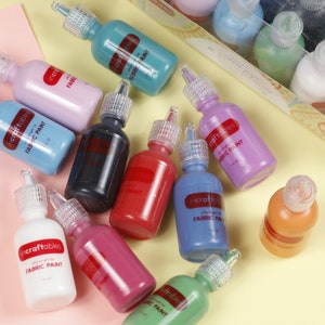 Może przedstawiać: Zestaw dziesięciu 1 fl oz butelek farby do tkanin w r&oacute;żnych kolorach, w tym r&oacute;żowym, niebieskim, zielonym, czerwonym, pomarańczowym i białym. Butelki są oznaczone "incraftables ultra bright fabric paint".