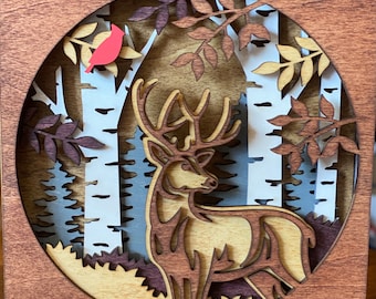 May include: A wooden wall art piece featuring a laser-cut design of a deer standing in a forest. The deer is brown and the forest is made up of trees with white bark and green leaves. A small red bird is perched on a branch in the top left corner of the image.