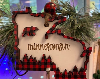 May include: Wooden Minnesota-shaped ornament with a red and black buffalo check border. The word "minnesconsin" is engraved on the white surface, with a bear and trees. A red and black plaid ball hangs from the top, attached with twine.