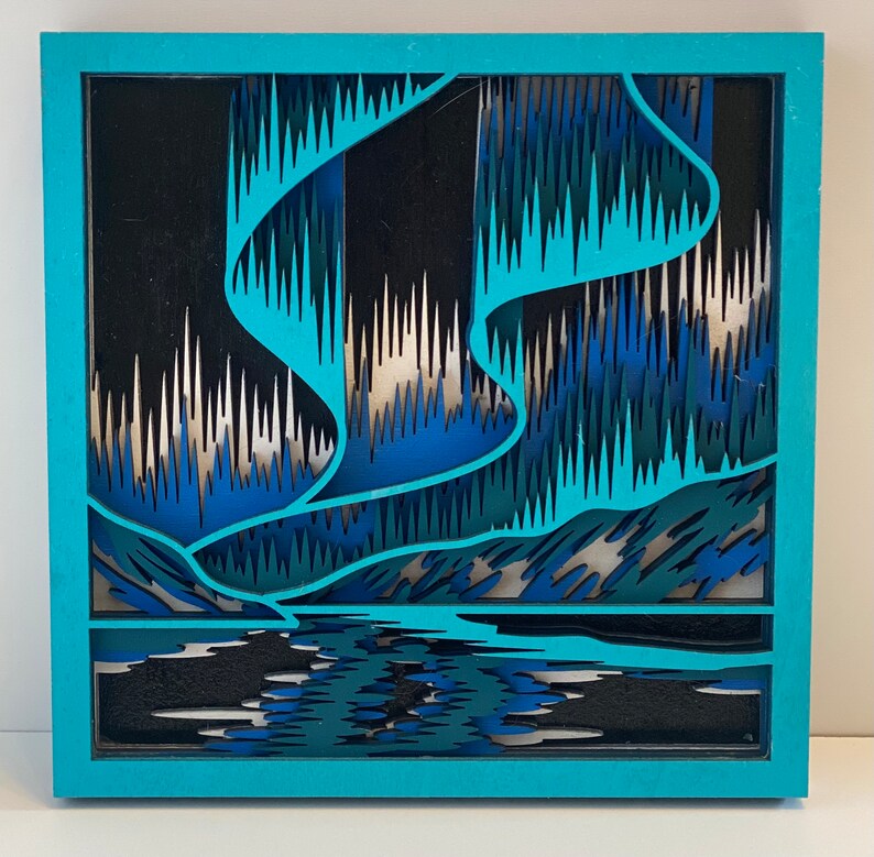 May include: A blue and green layered paper art piece depicting the aurora borealis, also known as the Northern Lights. The artwork is framed in a light blue frame.